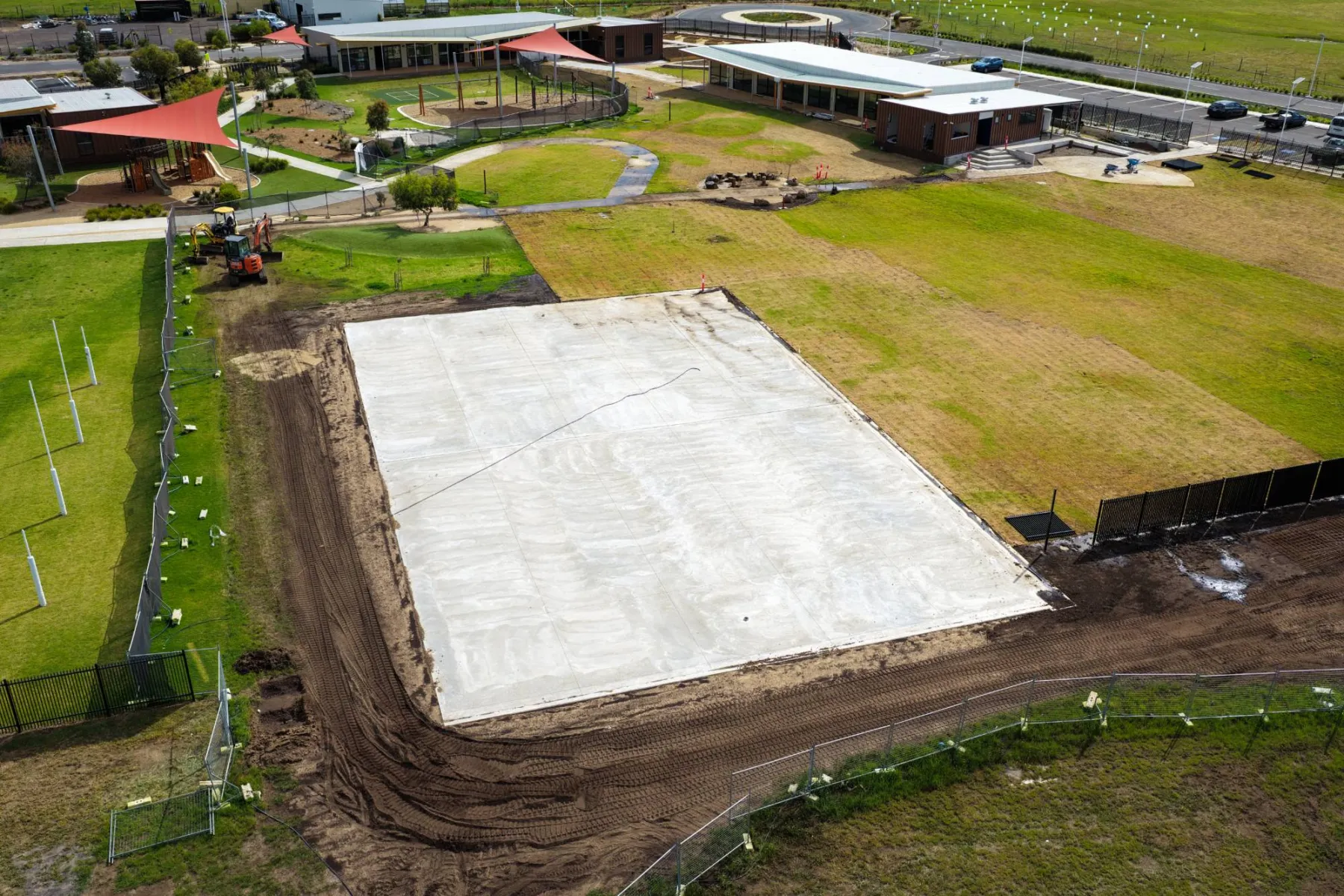 The freshly poured slab for the new sports court beside the new turf, with an excavator working the clean fill along the fence line